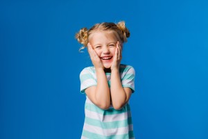 Cute little girl blonde smiling holding hands on sides standing on a blue background, happy child on a blue background, space for text